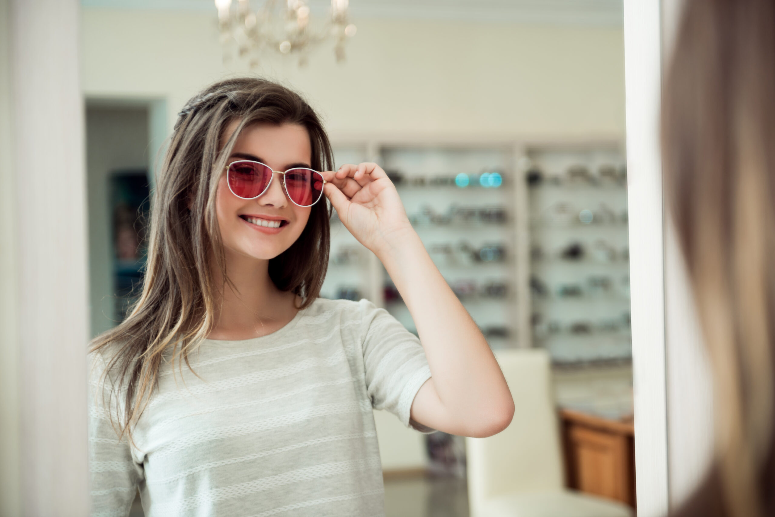 Stylish sunglasses make person look fashionable. Portrait of smiling good-looking young european girl in optician store, posing in mirror while trying on sunglasses, deciding which pair to buy.